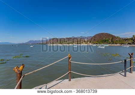 Fence With Wooden Posts And Ropes On The Promenade With Lake Chapala With Boats, The Town, The Pier 
