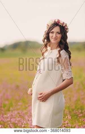 Pregnant Happy Girl Stand And Hold Hands On Stomach, Stand On Grass In The Outdoor In The Field Of P
