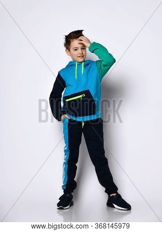 Cute Little Schoolboy With Stylish Hairdo In Bright Cotton Sportsuit Stands Smiling, Putting Hand Up