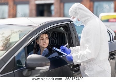 medicine, quarantine and pandemic concept - doctor or healthcare worker in protective gear or hazmat suit, medical mask, gloves and goggles with clipboard and woman in car waiting for coronavirus test