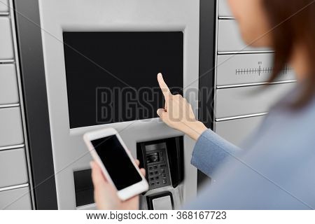 mail delivery and post service concept - close up of woman with smartphone choosing operation on outdoor automated parcel machine's touch screen
