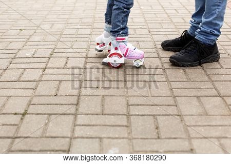 Dad Teaches His Little Daughter To Ride Four-wheeled Roller Skates. The Father Supports The Child By