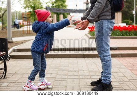 Dad Teaches His Little Daughter To Ride Four-wheeled Roller Skates. The Father Supports The Child By