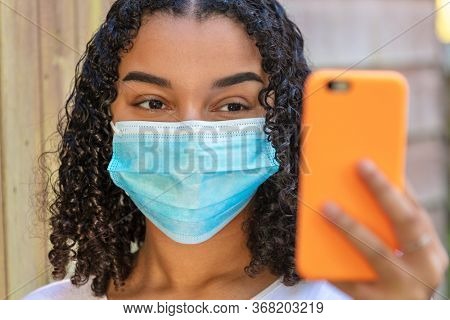 Mixed race African American teenager teen girl young woman wearing a face mask and using a mobile cell phone for social media or selfies during the Coronavirus COVID-19 pandemic
