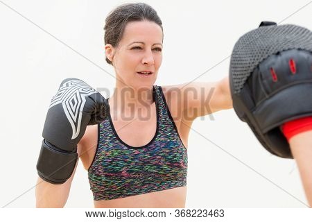 Concentrated Woman Punching Boxing Paw. Middle-aged Lady In Sport Bra Training Punch In Boxing Glove