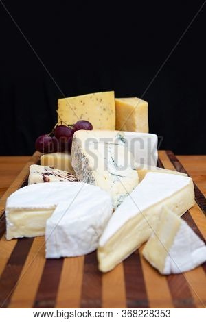 Heap Of Soft, Blue And Cheeses On Wooden Chopping Board Against Dark Wall Background. Closeup, Verti