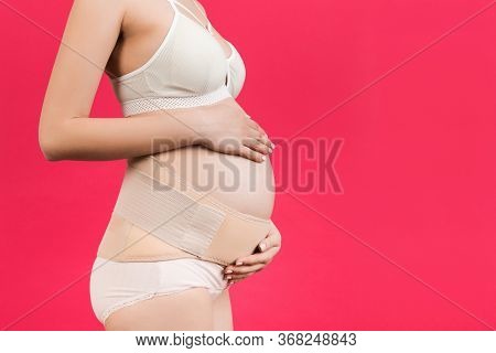 Side View Of Pregnant Woman In Underwear Wearing Pregnancy Bandage At Pink Background With Copy Spac