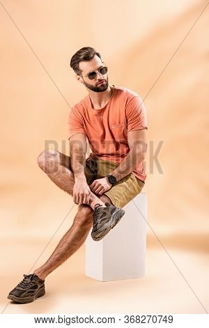 Stylish Man In Shorts, Summer T-shirt And Sunglasses Sitting On White Cube On Beige