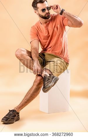 Handsome Young Man In Shorts, Summer T-shirt And Sunglasses Sitting On White Cube On Beige