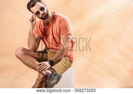 Fashionable Handsome Young Man In Shorts, Summer T-shirt And Sunglasses Sitting On White Cube On Bei