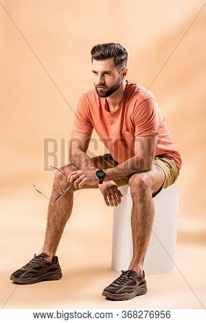 Young Bearded Man In Shorts And Summer T-shirt Holding Sunglasses Sitting On White Cube On Beige