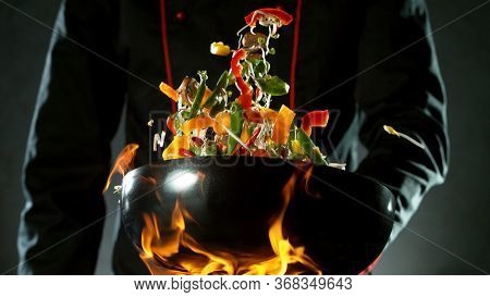 Closeup of chef throwing vegetable mix from wok pan in fire. Fresh asian food preparation on dark background.