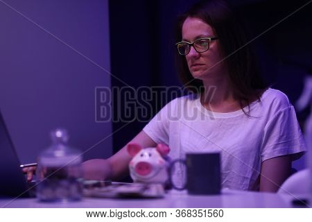 Portrait Of Upset Woman Sitting At Night. Glass Container With Coins. Depressed Female Calculating F