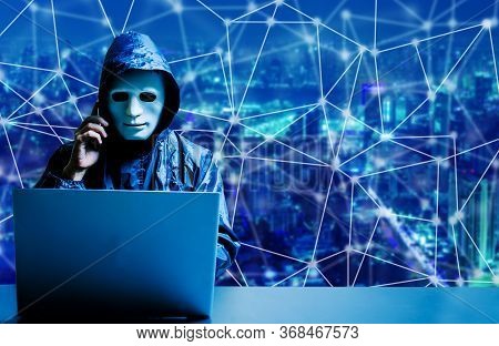 Anonymous Computer Hacker In White Mask And Hoodie. Obscured Dark Face Using Laptop Computer For Cyb