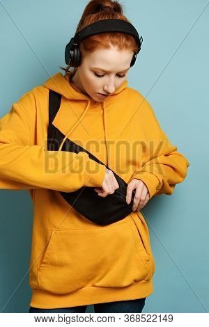 Red-haired Girl In A Yellow Sweatshirt And Headphones Stands On A Blue Background, Buttoning A Bag O