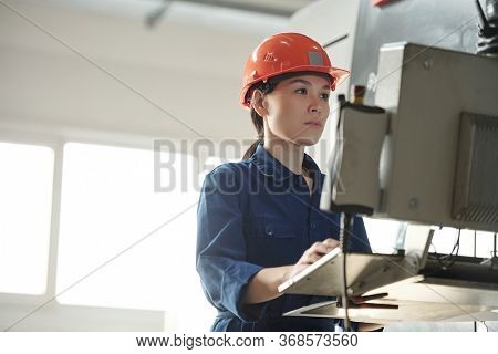Serious young Asian operator in hardhat using computer of industrial machine while choosing setups in workshop