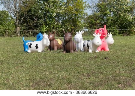 A Herd Of Inflatable Hopper Toys In The Field. Green With Yellow Flowers Pasture, Trees In The Backg