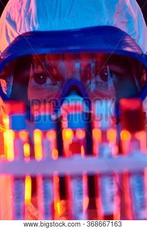 COVID-19. Close-up portrait of a doctor in a protective suit showing blood samples of infected people in test-tubes. Coronavirus test. Black background with red and blue light.