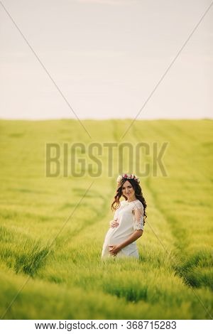Young Pregnant Woman In Fieid Of Green Grass. Young Beautiful Pregnant Girl With A Wreath On Her Hea