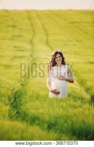 Young Pregnant Woman In Fieid Of Green Grass. Young Beautiful Pregnant Girl With A Wreath On Her Hea