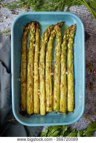 Roasted asparagus seasoned with salt, pepper, garlic and decorated with fresh herbs. Served in blue heatproof dish. Placed on gray stone background.