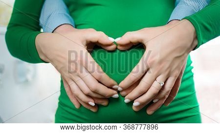 Pregnant Belly With Fingers Heart Symbol. Beautiful Young Pregnant Woman And Her Husband Together Ca