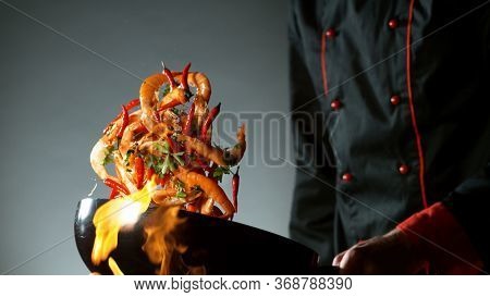 Closeup of chef throwing prawns from wok pan in fire. Fresh asian food preparation on dark background.
