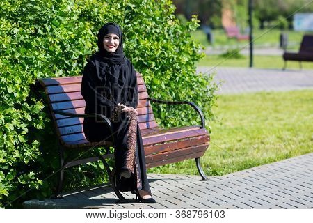 A Beautiful Young Muslim Woman Is Sitting On A Bench. Islamic Girl Smiles Broadly, Resting On A Park