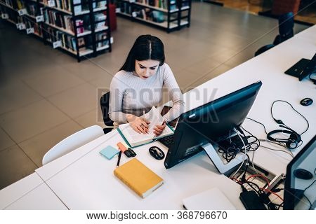 Insightful Female Student Reading Textbook And Using Computer While Studying In Library