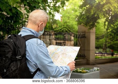 The Gray-haired Man In A Denim Shirt And With A Backpack Looks At The Map.