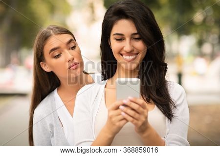 Jealous Friend. Envious Girl Peeking At Girlfriends Phone While She Texting Walking Together Outdoor