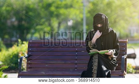 Muslim Woman Is Reading A Book On A Bench. Islamic Girl In National Dress Is Sitting With Book In He