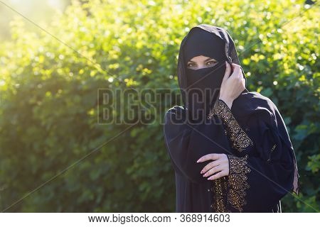 Beautiful Muslim Woman On A Background Of Green Leaves. Islamic Woman In Oriental Clothes Stands Aga
