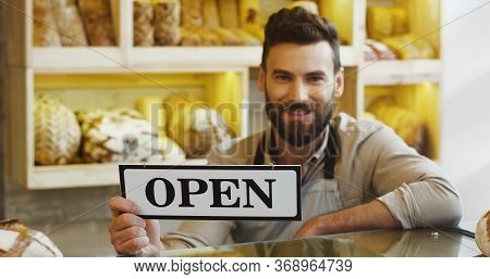 Portrait Of Handsome Smiling Baker Holding An Open Signboard In The Bakery Shop.