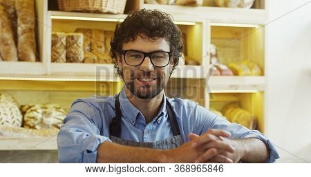 Close Up Of The Smiled Attractive Male Seller In Glasses Posing To The Camera In The Bakery Shop At 