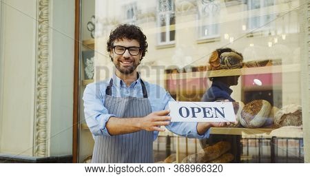 Male Caucasian Smiled Bakery Vendor Putting And Turning Signboard Open On The Glass Door Of The Shop