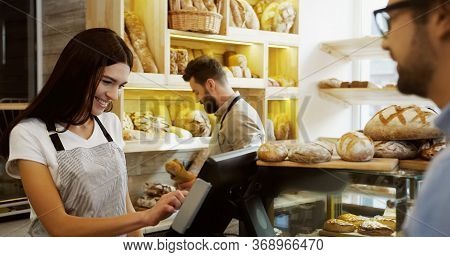 Young Beautiful Woman Standing At The Counter In The Bakery Shop And Selling Bread To The Man, Her M