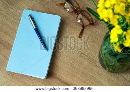 A Blue Notebook With A Ballpoint Pen Lies On A Table Next To A Vase With Rapeseed Flowers