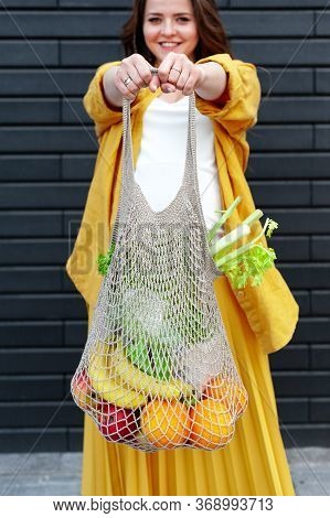 Pregnant Woman In Yellow Clothes With An Eco Bag With Fruits And Vegetables On A Black Wall Backgrou