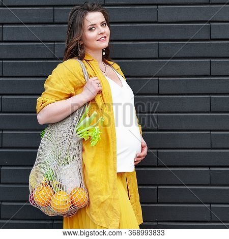 Pregnant Woman In Yellow Clothes With An Eco Bag With Fruits And Vegetables On A Black Wall Backgrou