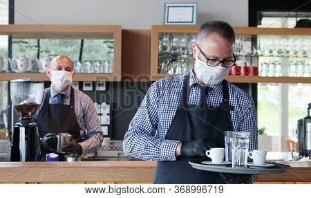 waiter in a medical protective mask serves  the coffee in restaurant durin coronavirus pandemic representing new normal concept