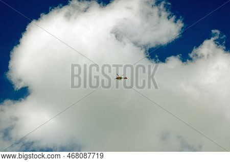 View From The Ground Of The London Air Ambulance Flying Overhead On A Sunny Day With White Fluffy Cl