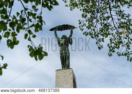The Historic Liberty Statue Upon Gellert Hill In Front Of A Cloudy Sky And Green Leaves In Budapest,