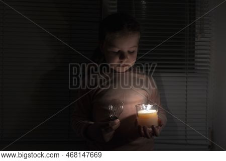 A Girl In A Dark Room Holds A Candle And An Electric Light Bulb In Her Hands. Power Outage Concept. 