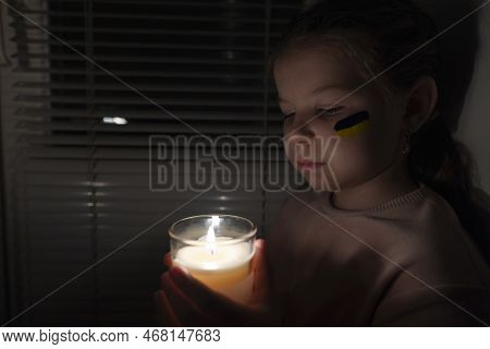 A Girl With A Painted Flag Of Ukraine On Her Cheek In A Dark Room Holds A Candle In Her Hands. Power