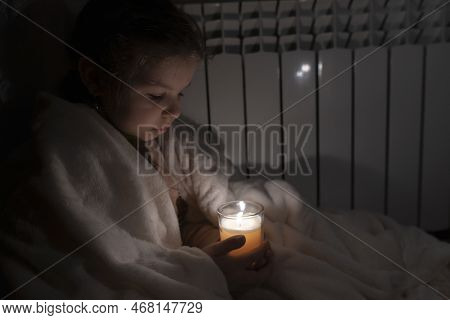 Girl In A White Plaid Holds A Candle In A Glass Cup In Her Hands Against The Background Of A Househo