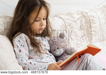 Unhappy Little Girl In Sleepwear Sitting On Couch And Reading Book. Portrait Of Child, Preschooler. 
