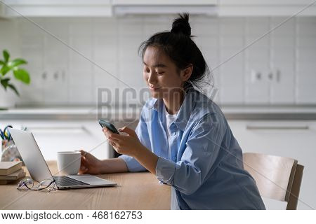 Smiling Asian Girl Freelance Worker Sitting At Workplace Holding Smartphone Receiving Message Chatti