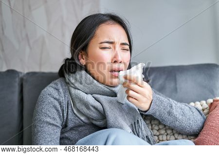 Close Up Of Korean Girl Sneezing In Napking, Sitting At Home In Scarf, Feeling Unwell, Concept Of Wi