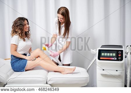 Young Cosmetologist Performing Procedure Of Laser Removal Of Unwanted Hair On Legs Of Female Client.